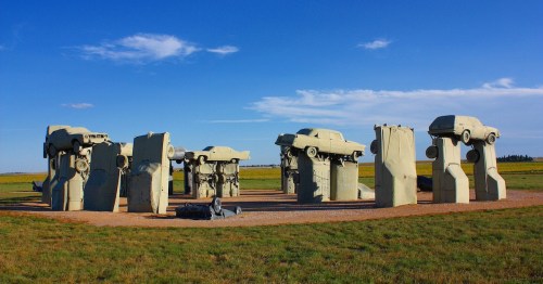 A unique sculpture installation featuring cars mounted on stone-like structures in an open field under a blue sky.