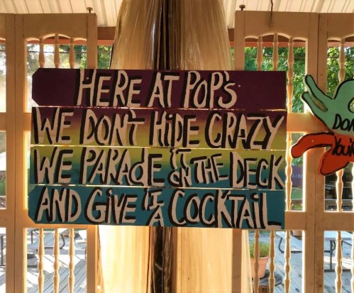 Colorful wooden sign with text: "Here at Pop's, we don't hide crazy, we parade on the deck and give it a cocktail."