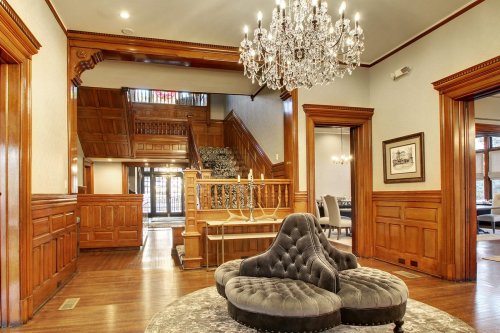 Elegant foyer with a chandelier, wooden paneling, and a plush gray seating area, leading to a grand staircase.