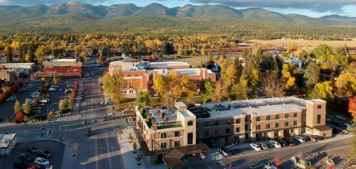 Aerial view of a town with colorful autumn trees, mountains in the background, and a mix of buildings and parked cars.