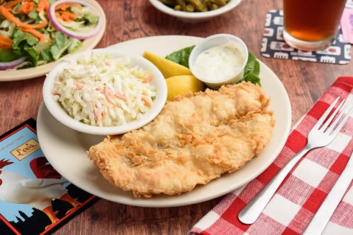 A plate of fried fish with coleslaw, a lemon wedge, and a side salad, set on a wooden table with a drink.