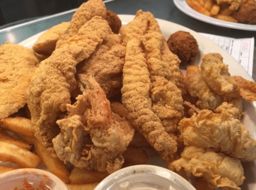 A plate of fried fish fillets, shrimp, hushpuppies, and French fries, served with a side of dipping sauce.