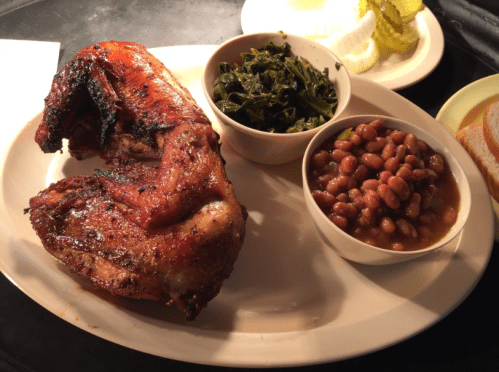 A plate featuring grilled chicken, collard greens, baked beans, and a side of pickles and bread.
