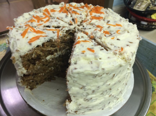 A layered carrot cake with cream cheese frosting and orange carrot shavings, partially sliced on a silver platter.