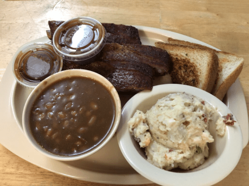 A plate with BBQ ribs, two sauces, baked beans, potato salad, and toasted bread.