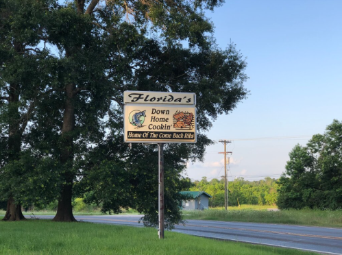 Sign for "Florida's Down Home Cookin'" featuring a fish and a barbecue graphic, with a rural landscape in the background.