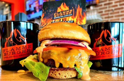 A delicious Flamme Burger with melted cheese, fresh veggies, and two branded mugs in the background.