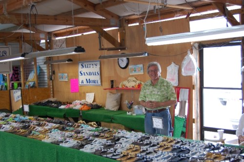 A smiling vendor stands behind a table filled with various sandals and shoes in a market setting.