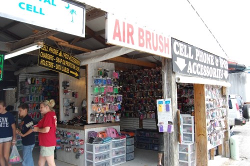 A colorful outdoor market stall displaying cell phone accessories and airbrush items, with shoppers browsing.