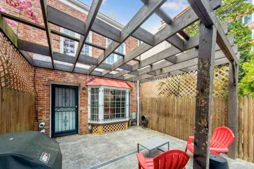 A cozy outdoor patio with a wooden pergola, brick walls, and red chairs, featuring a grill and a small table.