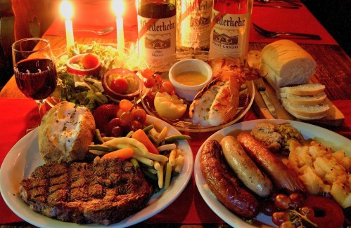 A beautifully arranged dinner table featuring steak, sausages, vegetables, bread, and wine, illuminated by candlelight.