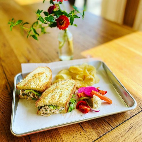 A toasted sandwich with greens, served with chips and a side of colorful pickled vegetables, on a metal tray.