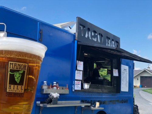 A taco truck with a sign reading "Taco Bar" and a pint of beer in the foreground, set in a sunny outdoor location.