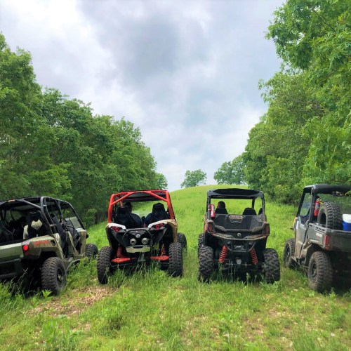 Four off-road vehicles parked on a grassy trail, surrounded by trees under a cloudy sky.