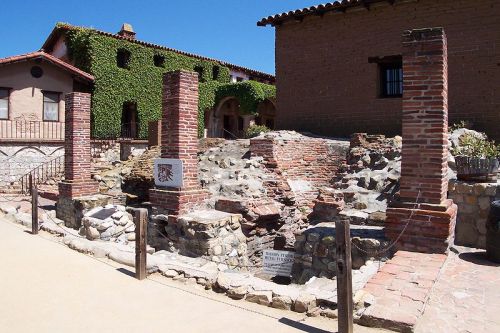 Ruins of a historic building with brick pillars and stone foundations, surrounded by greenery and a clear blue sky.