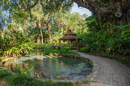 A serene garden scene featuring a gazebo by a pond, surrounded by lush greenery and trees.