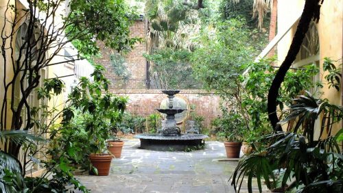A serene courtyard featuring a stone fountain surrounded by lush greenery and potted plants.