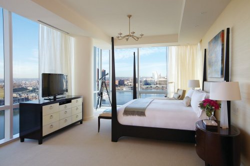 Modern bedroom with large windows, a bed, TV, and a view of the city skyline and water. Bright and airy atmosphere.