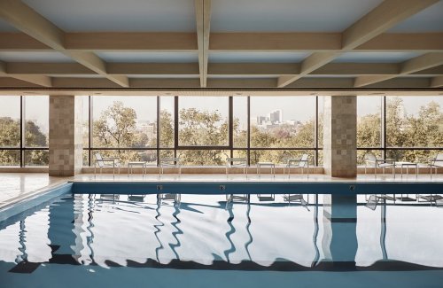 Indoor swimming pool with a calm water surface, large windows showcasing trees and a city skyline in the background.