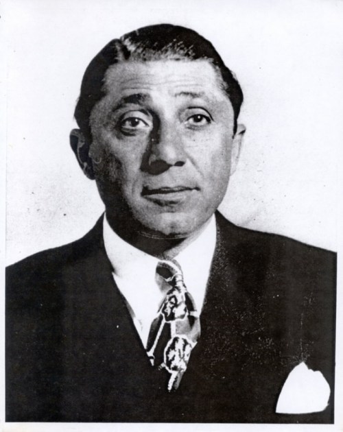 Black and white portrait of a man in a suit with a patterned tie, looking directly at the camera.