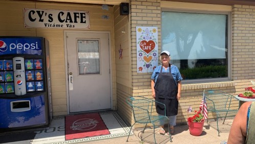 A woman in an apron stands outside Cy's Cafe, next to a vending machine and a sign with hearts.