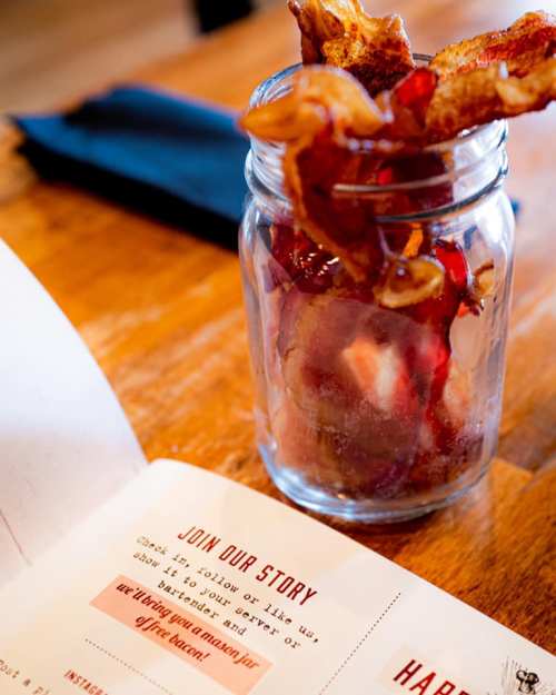 A jar filled with crispy bacon strips sits on a wooden table next to a menu.