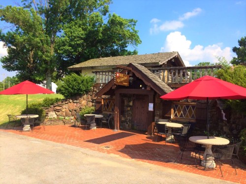 A charming stone building with a wooden sign, outdoor seating, and red umbrellas on a sunny day.