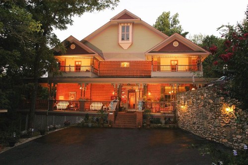 A charming two-story brick house with warm lights, surrounded by trees and a stone wall, creating a welcoming atmosphere.
