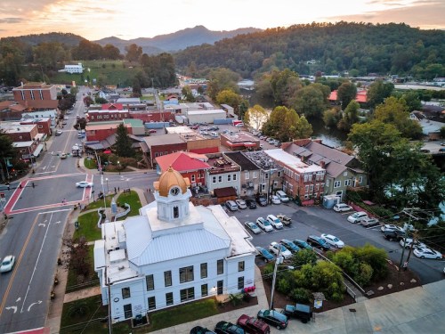 Aerial view of a small town with a clock tower, colorful buildings, and surrounding hills at sunset.