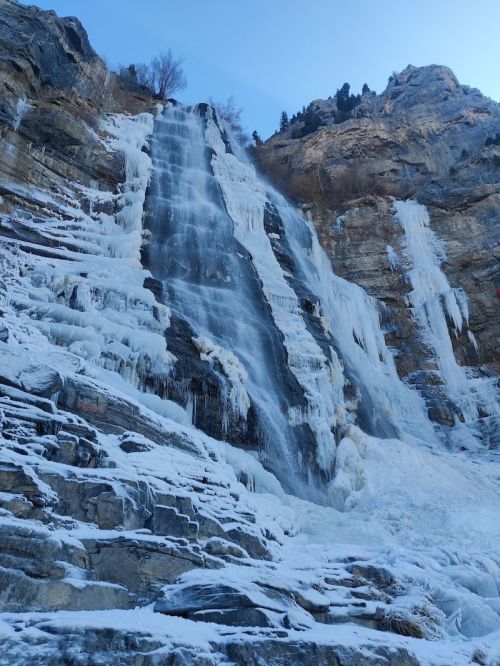 A frozen waterfall cascading down rocky cliffs, surrounded by icy formations and a clear blue sky.