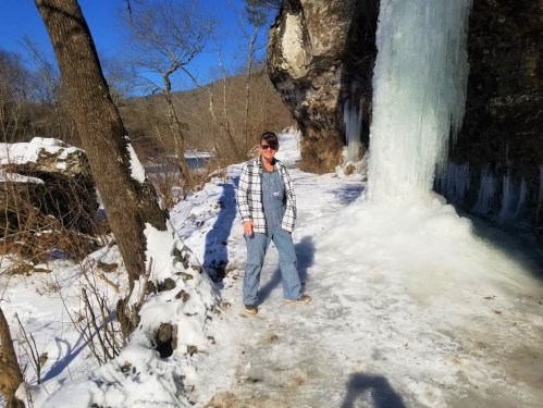 A person stands near a frozen waterfall, surrounded by snow and trees, under a clear blue sky.