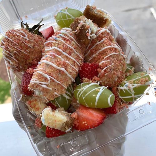 A clear container filled with strawberries, grapes, and dessert bites, drizzled with white chocolate and colorful sprinkles.