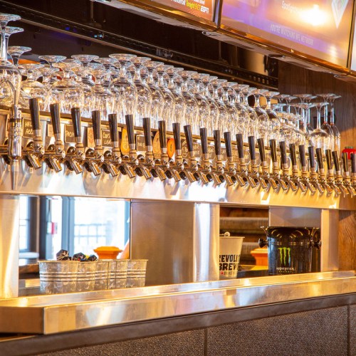 A bar with numerous beer taps and glasses lined up, featuring a modern design and screens above.