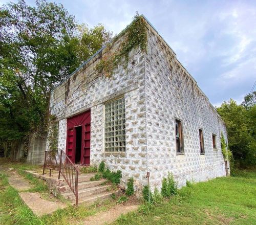 Abandoned building with a textured stone exterior, red double doors, and overgrown grass surrounding it.