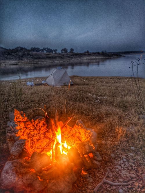 A glowing campfire in the foreground with a tent by a calm river at dusk, surrounded by grass and rocks.