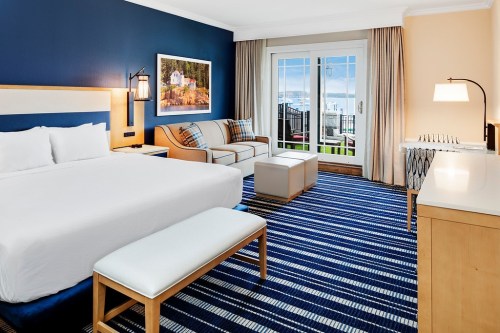 A modern hotel room featuring a king bed, seating area, and large windows with a view, decorated in blue and beige tones.