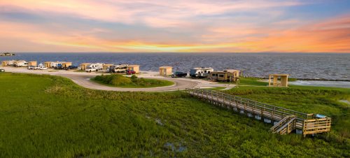 A scenic view of RVs parked by the water, with a wooden walkway and vibrant sunset sky in the background.