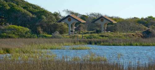Two pavilions with thatched roofs beside a calm water body, surrounded by lush greenery and tall grasses.