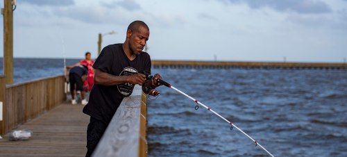 A man fishing from a wooden pier, focused on his rod, with water and a cloudy sky in the background.