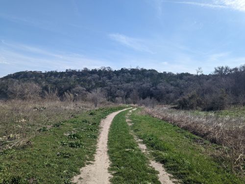 A winding dirt path leads through a grassy area, with a hillside covered in trees in the background under a clear blue sky.