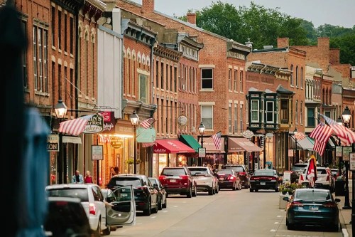 A charming street lined with brick buildings, shops, and American flags, bustling with parked cars and pedestrians.