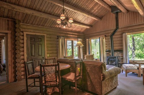 Cozy log cabin interior with a dining table, chairs, and a fireplace, surrounded by large windows and natural light.