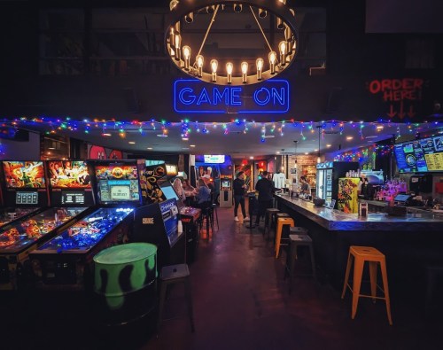 A vibrant arcade bar with pinball machines, colorful lights, and a lively atmosphere. "Game On" sign illuminated above.