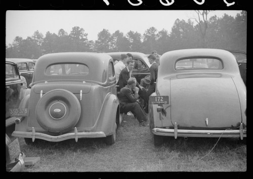 Two vintage cars parked closely together, with a group of people gathered nearby in a field.