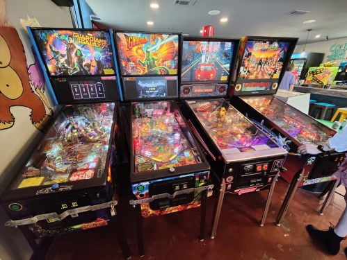 A row of colorful pinball machines with vibrant artwork and illuminated playfields in a lively arcade setting.