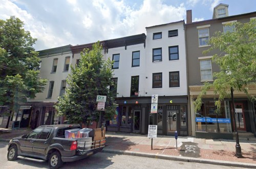 A street view of a row of buildings with trees, parked cars, and storefronts under a partly cloudy sky.