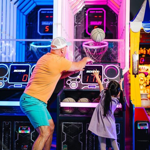 A man and a girl play basketball arcade games, with colorful lights and scoreboards in the background.