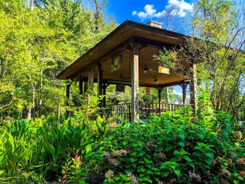 A wooden gazebo surrounded by lush greenery and colorful plants under a bright blue sky.