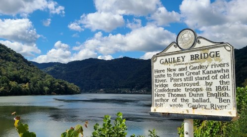 A historical sign by the river, detailing the union of New and Gauley rivers to form the Kanawha River, with mountains in the background.