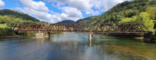 A rusty train bridge spans a calm river, surrounded by lush green hills and a partly cloudy sky.